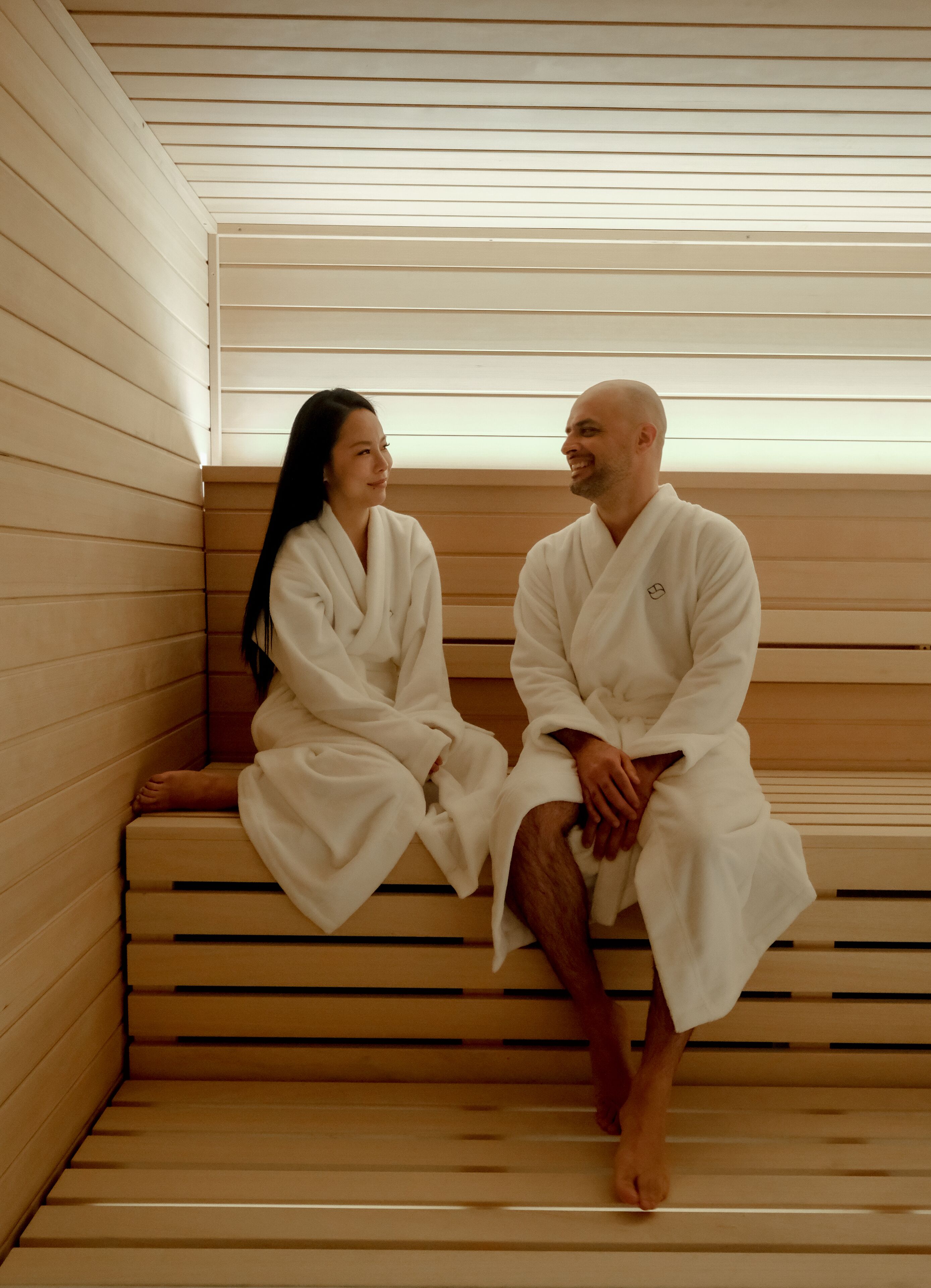 Two people in white robes sitting on wooden benches in a sauna.