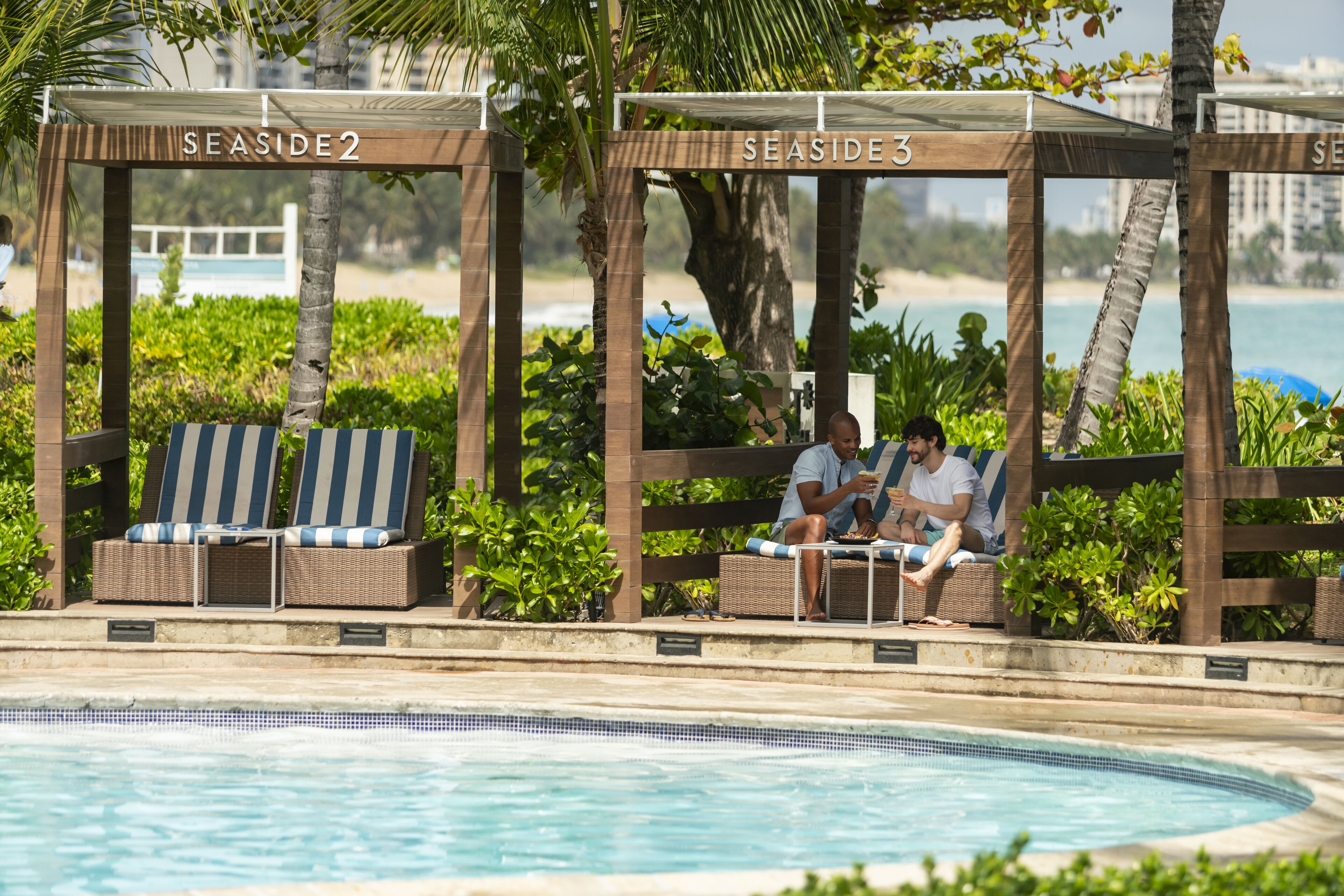 Un área junto a la piscina con sillones de descanso debajo de pérgolas de madera identificadas como "SEASIDE 2" y "SEASIDE 3".