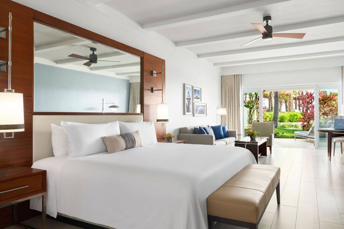 A hotel room with a large bed, a desk, a chair, a bench, and a balcony with a view of greenery.