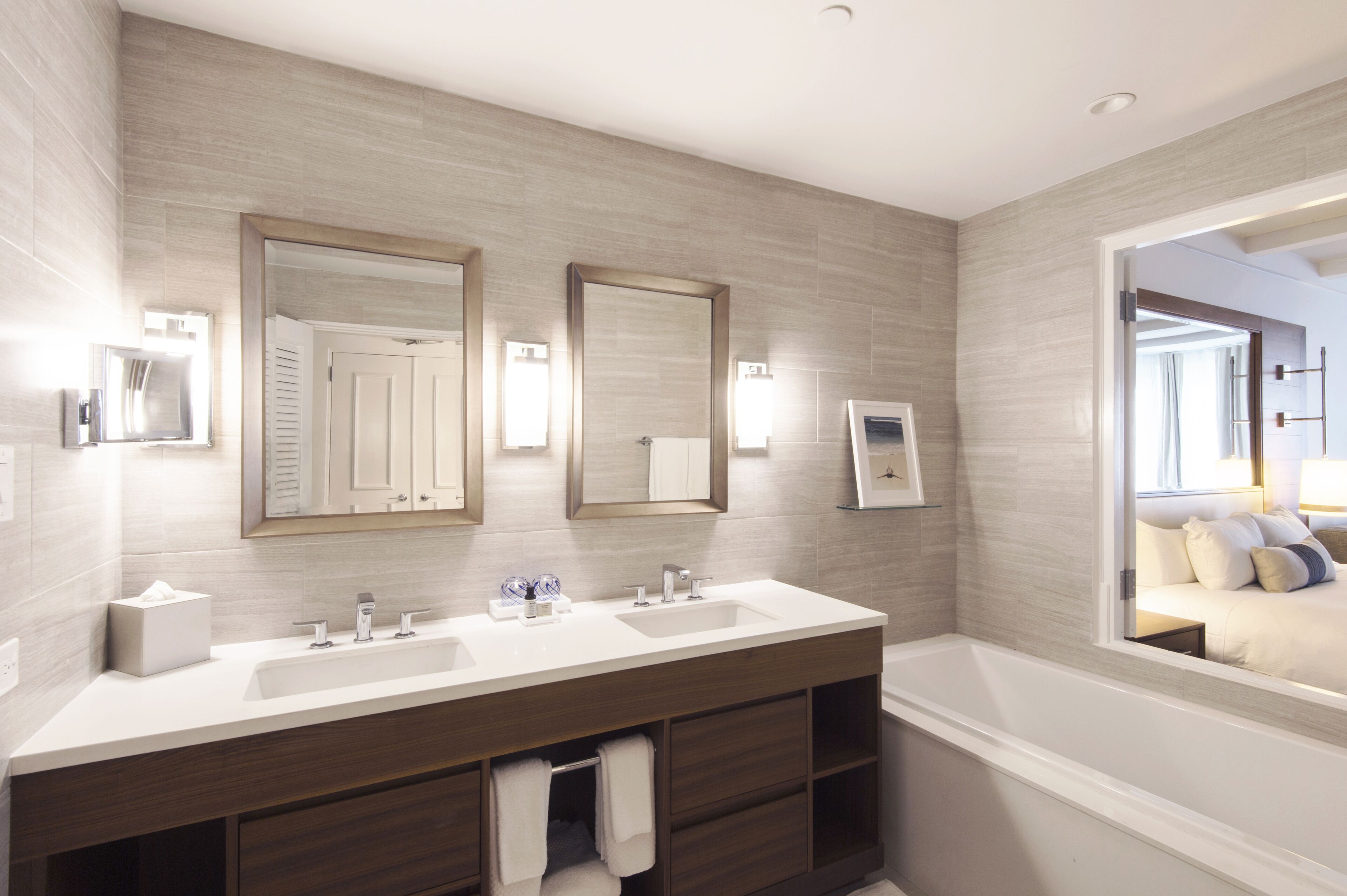 A modern bathroom with two sinks, a large mirror, and a view of a bedroom.