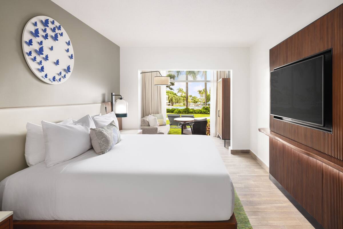 A modern hotel room with a large bed, a flat-screen TV, a view of the outdoor pool area, and a decorative butterfly wall art piece.