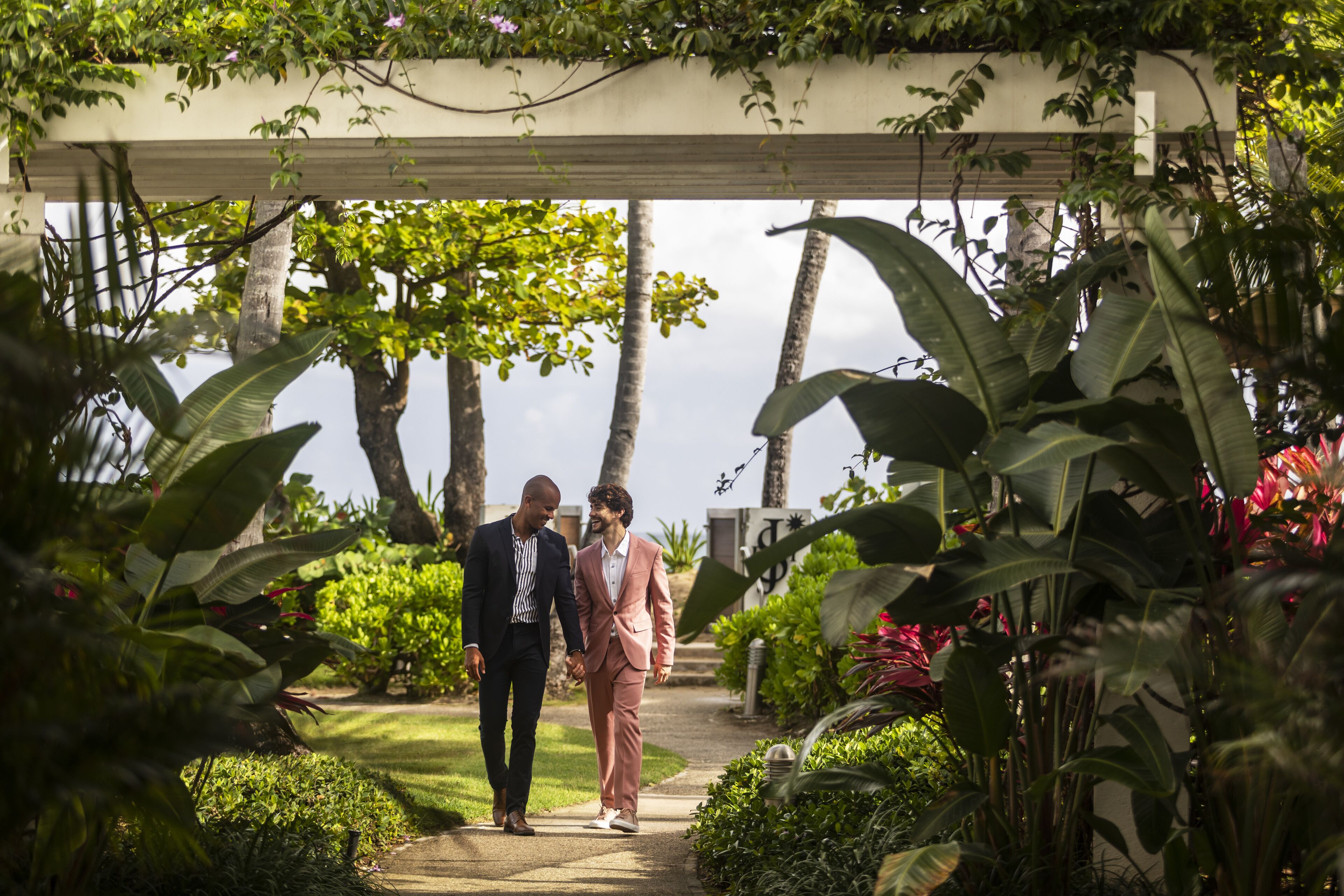 A couple walking under a covered pathway surrounded by lush greenery and tropical plants.