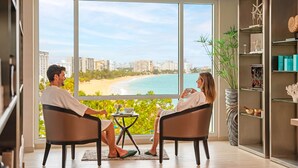A room with a large window overlooking a beach, a bookshelf, and a small table with a bowl and a plant. - Fairmont El San Juan Villa Experience (Carolina)