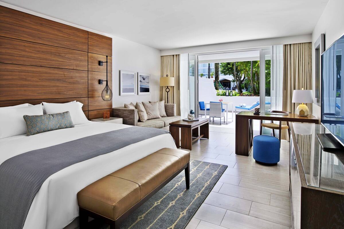 A modern hotel room with a large bed, a sofa, a desk, and a view of the outdoor pool area.