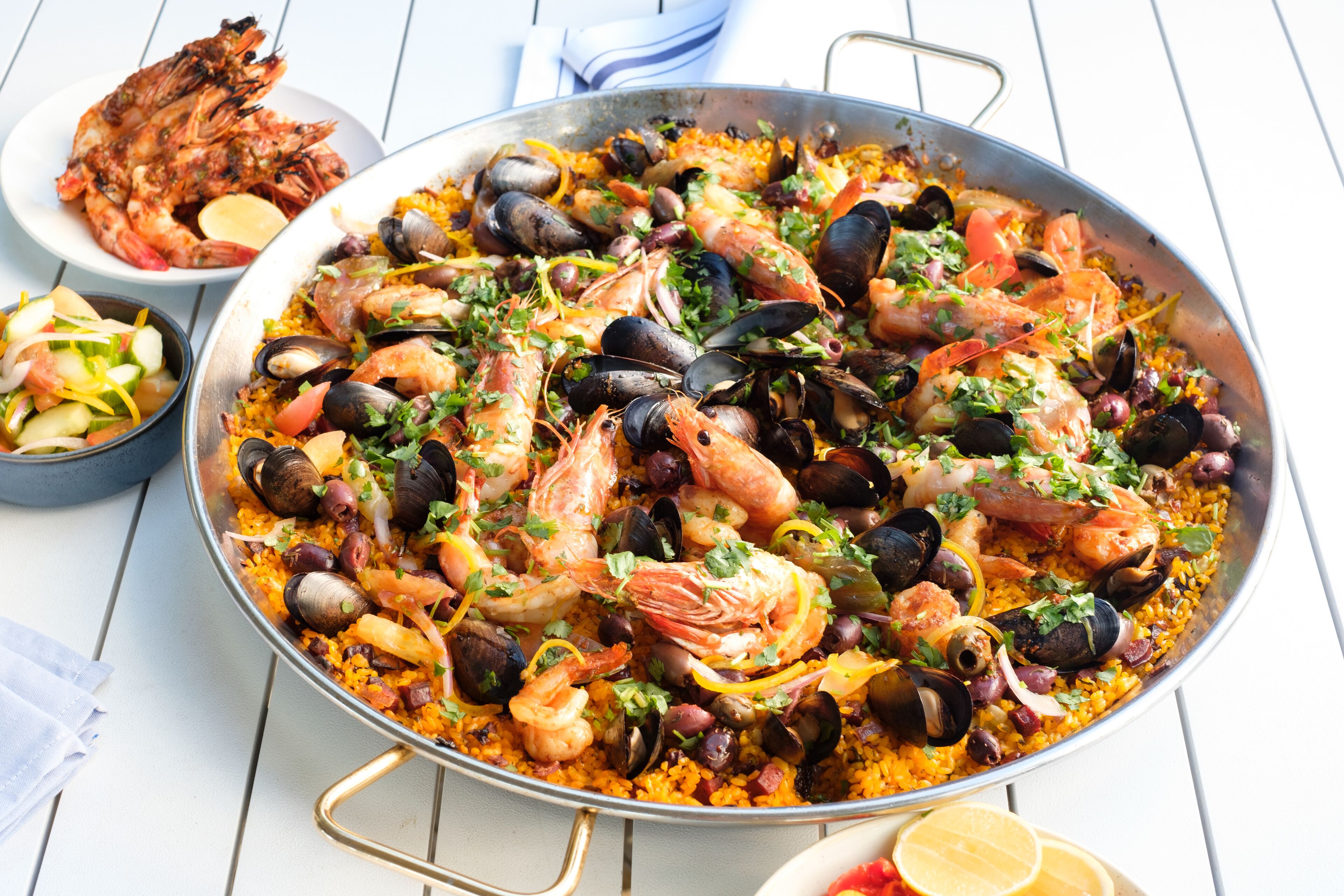 Eine große Paella mit Meeresfrüchten, darunter Garnelen und Muscheln, sowie Reis, dazu eine Schüssel gegrillte Garnelen und ein kleiner Salat.