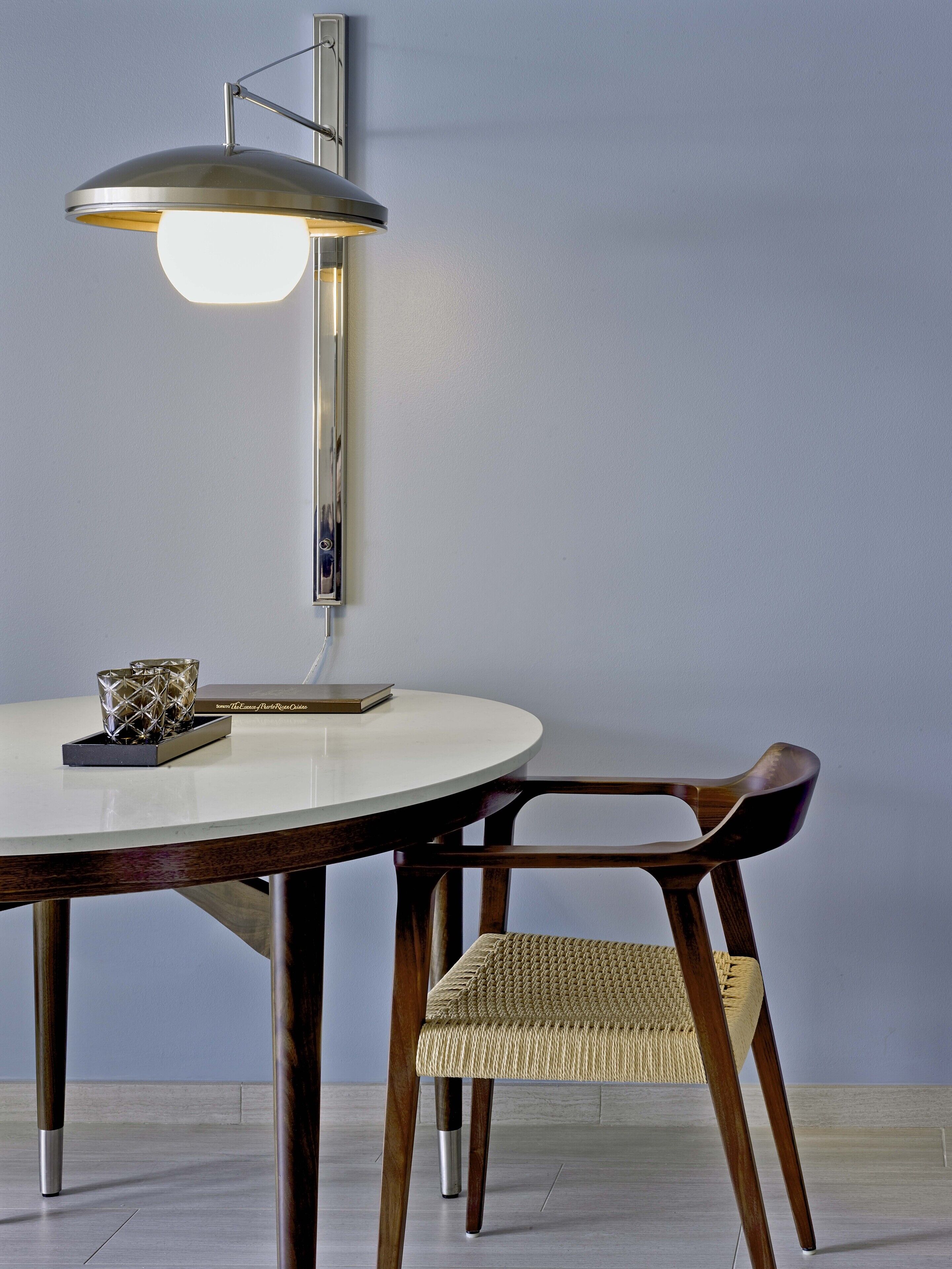 A round dining table with a white top, a dark wooden chair with a woven seat, and a wall-mounted lamp with a metallic shade.