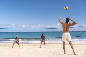 Un partido de voleibol de playa con jugadores en una cancha arenosa, una red y el océano al fondo.