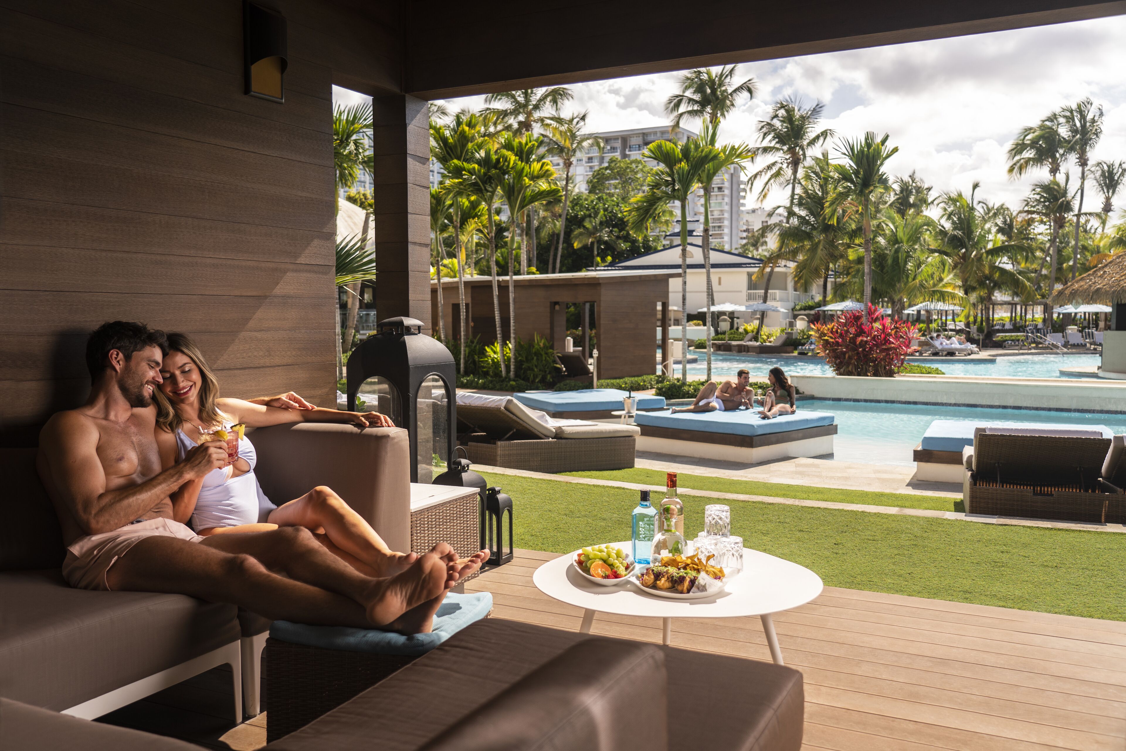 A couple relaxing on a sun lounger by a poolside, with a dining table and chairs nearby.
