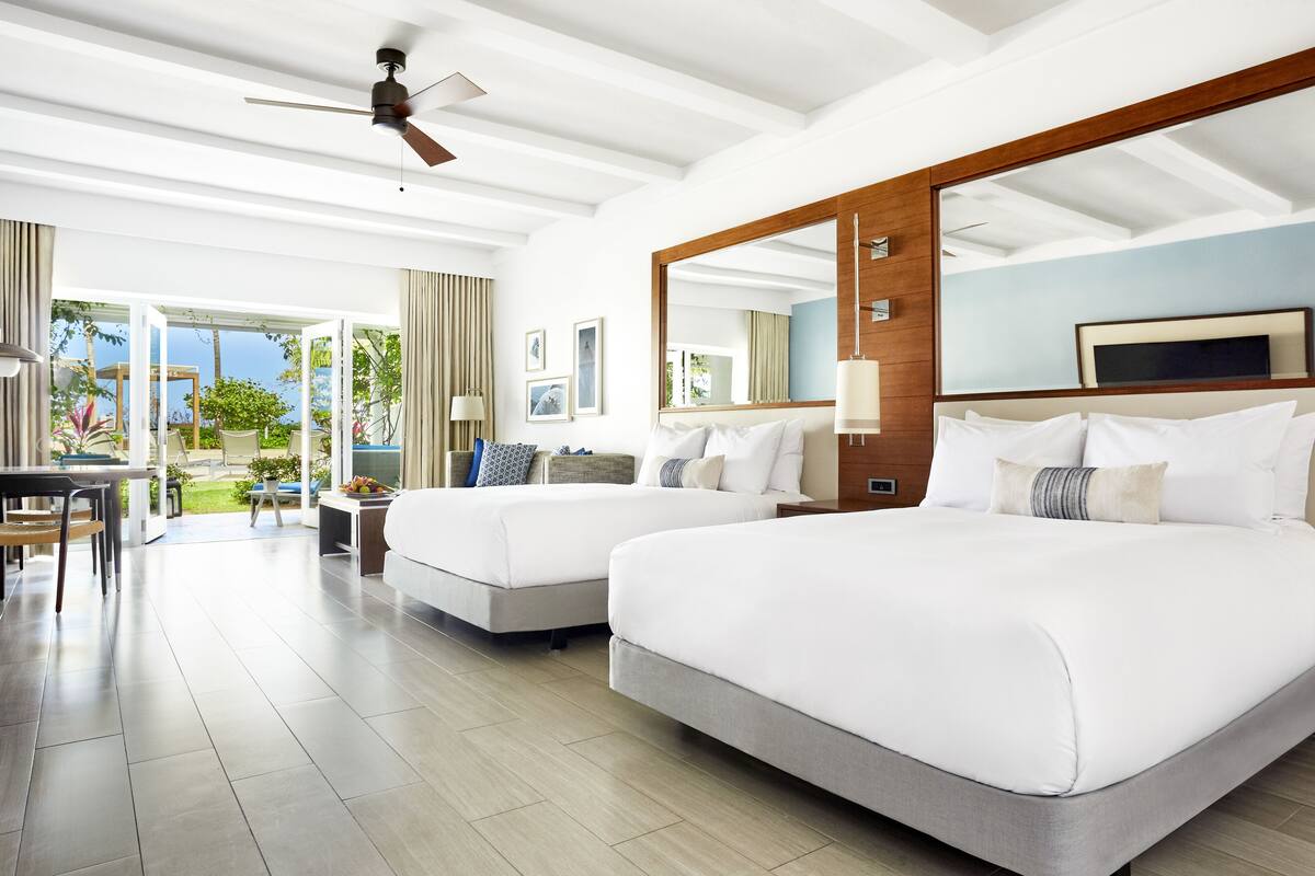 A modern hotel room with two beds, a large window, and a ceiling fan.