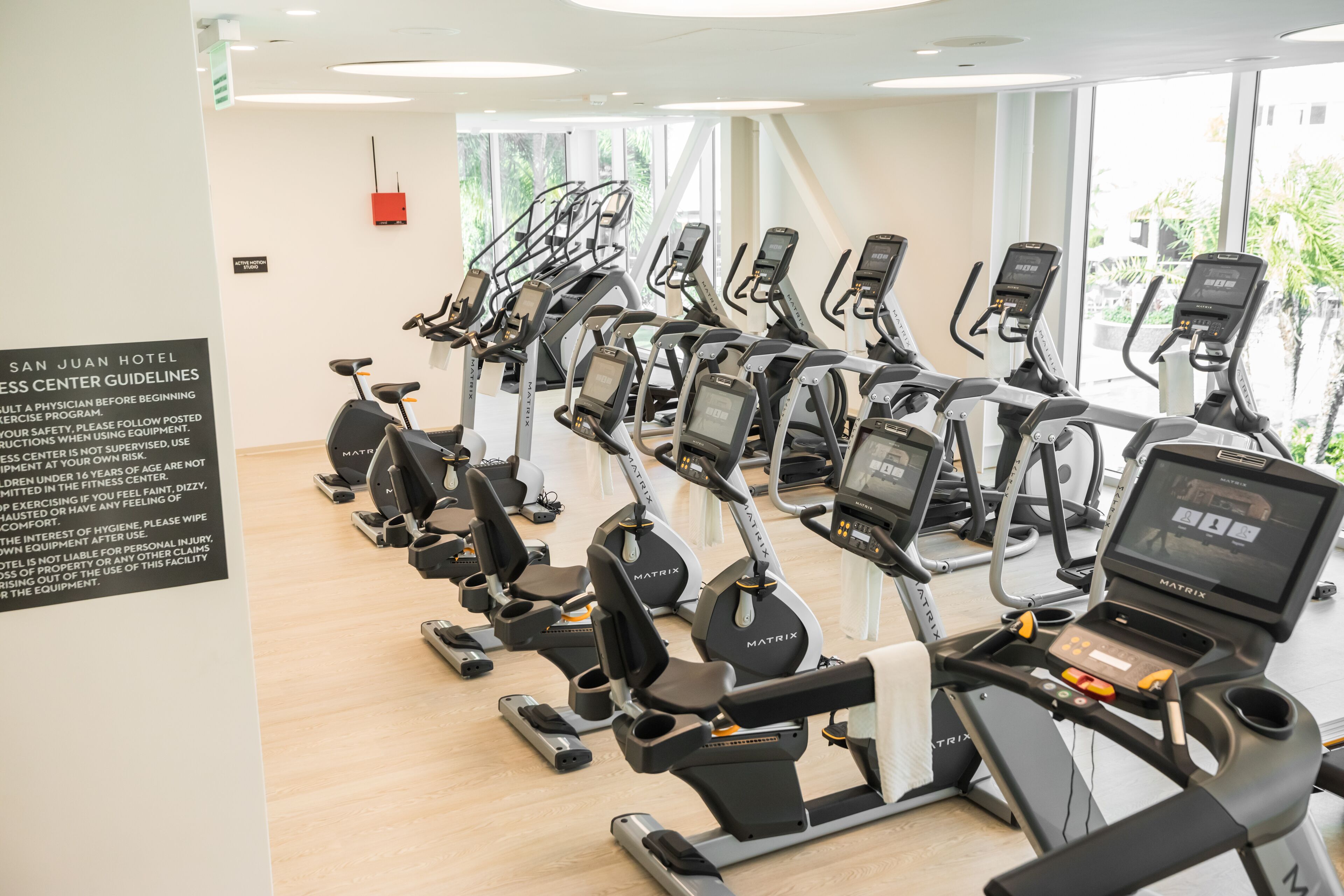 A row of stationary bikes in a well-lit room with large windows, a sign with guidelines, and a red emergency phone.