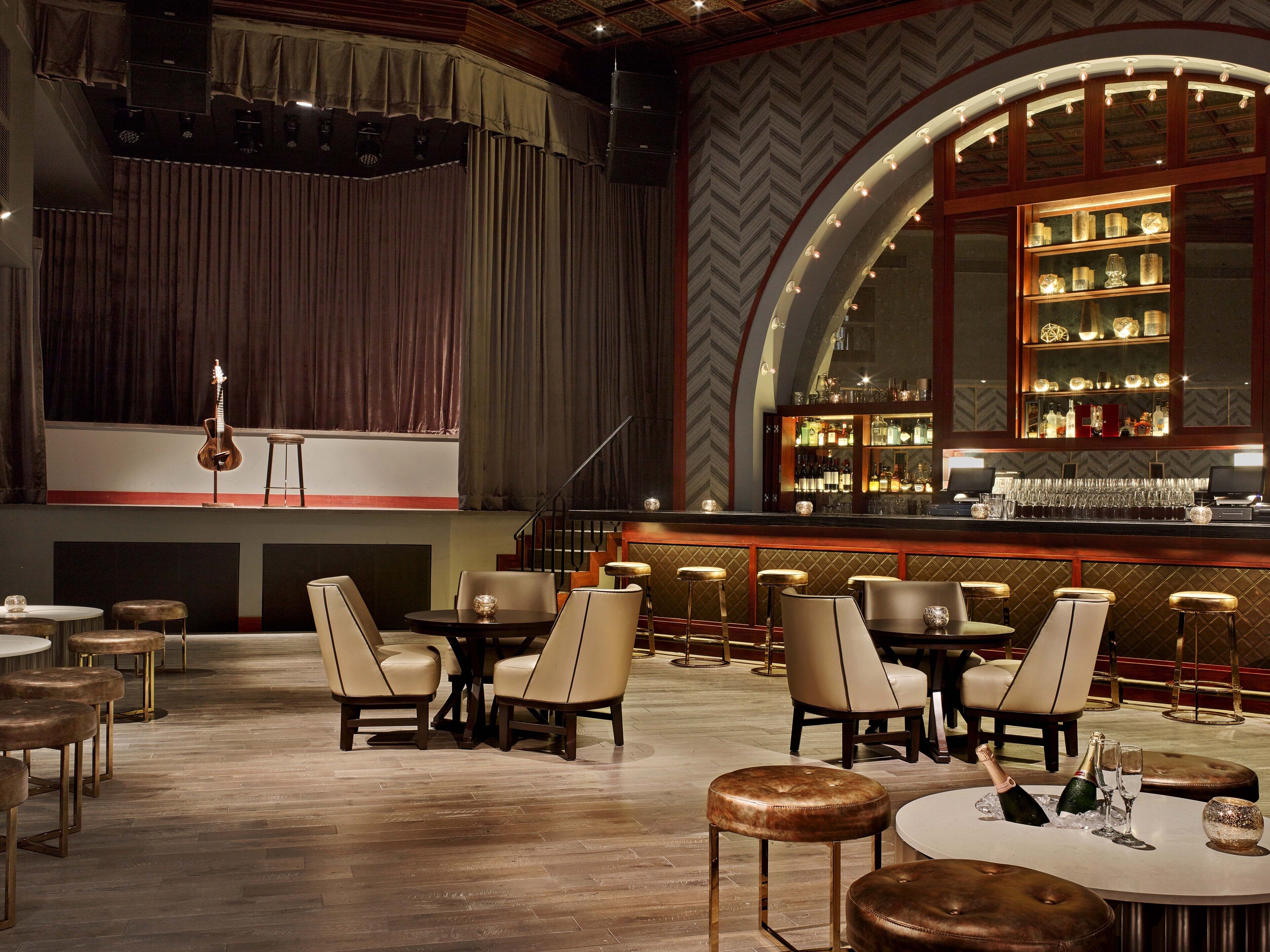 Zona de bar con escenario, taburetes y un área de estar con mesas y sillas.