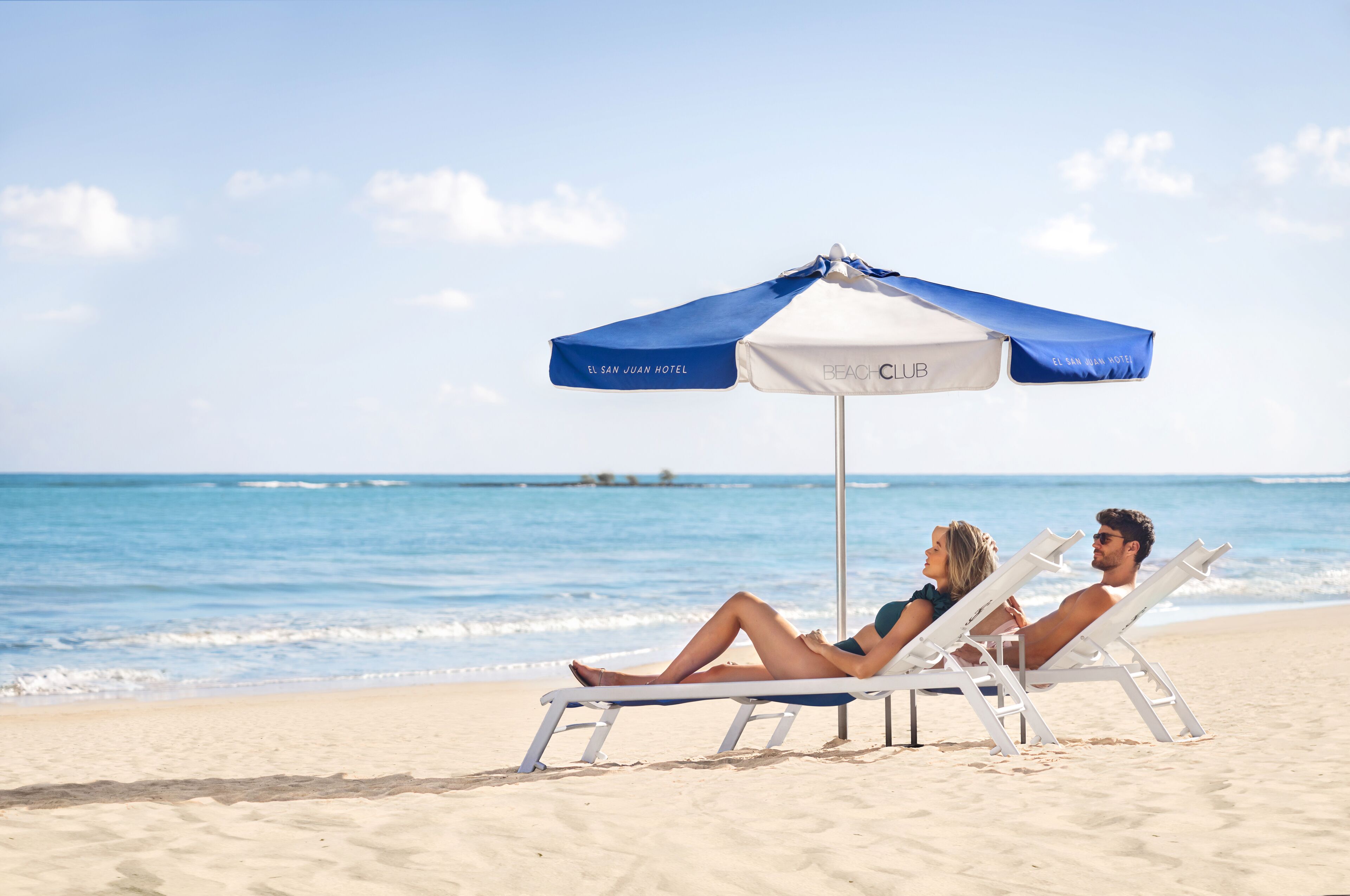 Two people relaxing on beach chairs under a beach umbrella at a hotel.