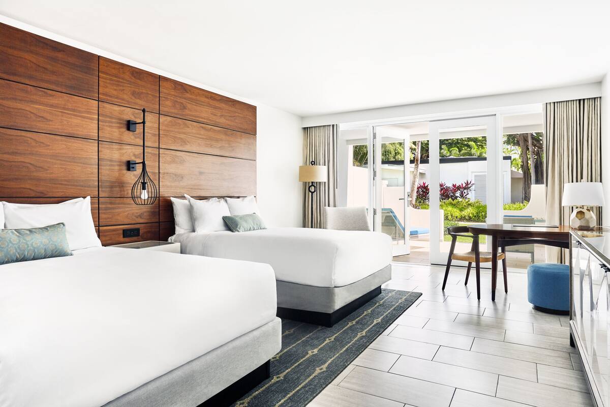 A hotel room with two beds, a wooden headboard, a dining area with a table and chairs, and a view of the outdoors.