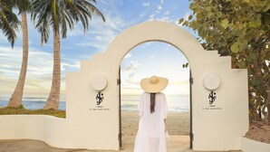 A woman in a white dress and hat walking through an arched doorway with palm trees and a beach view. - Fairmont El San Juan Villa Experience (Carolina)
