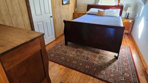 Une chambre avec un plancher en bois, un lit avec une tête de lit, une table de chevet, une commode en bois et une fenêtre avec des rideaux.
