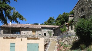 A rustic stone building with a tiled roof, a staircase leading to a higher level, and a small garden with pink flowers. - Gite 6 people in a stone farmhouse, swimming pool, property 10Ha, panoramic view (Rocles)