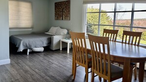 A room with a bed, a small table, a dining table with chairs, a chandelier, and a large window with a view.