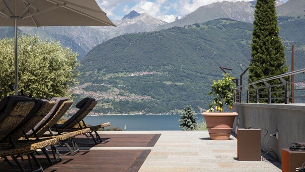 A terrace with wicker lounge chairs, a potted plant, and a view of a lake and mountains.