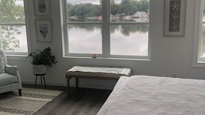 Un dormitorio con un ventanal grande que da a un lago, una cama, un banco y una silla.