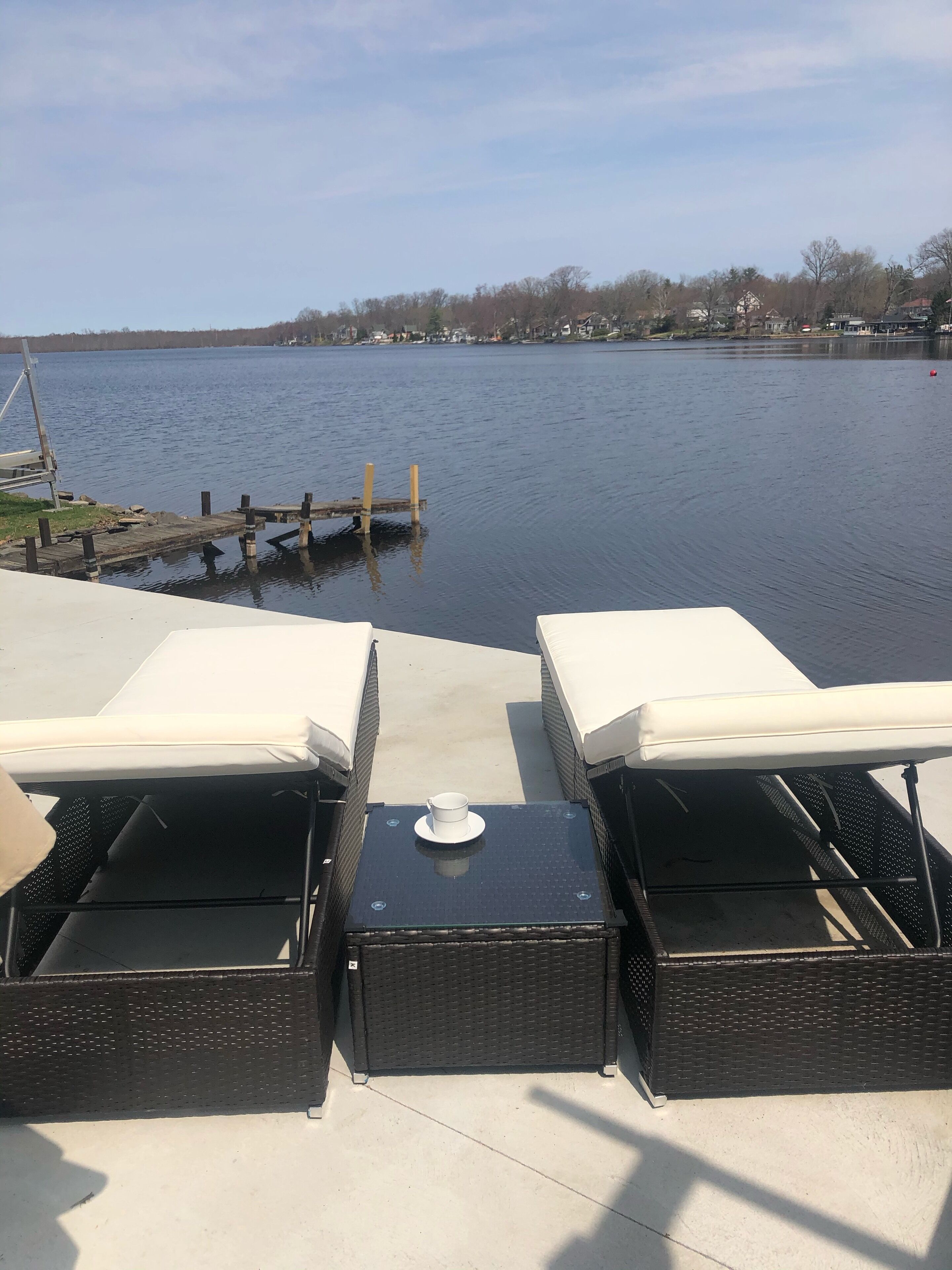 Uitzicht vanaf een terras met twee ligstoelen en een tafel, uitkijkend op een rustig meer met een steiger en gebouwen in de verte.
