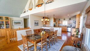 Un comedor amplio con una gran mesa de madera y sillas, una cocina al fondo y una estantería llena de libros.