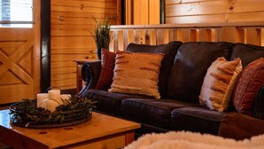 Interior acogedor de una cabaña con techo de madera, un sofá de cuero con cojines, una mesa de centro con vela y plantas, y una puerta de madera.