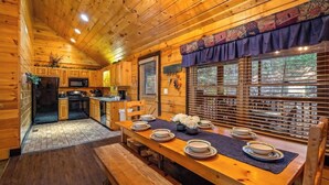 Interior de una cabaña de madera con una mesa de comedor preparada para cuatro, zona de cocina con electrodomésticos y ventanas grandes con persianas.