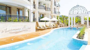 A swimming pool with lounge chairs and umbrellas, a gazebo, and a building with balconies.