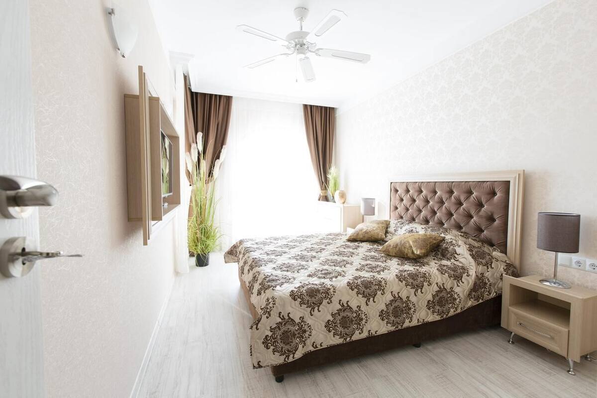 A bedroom with a tufted headboard, a bed with patterned bedding, a bedside table with a lamp, and a ceiling fan.