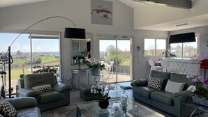 A spacious living room with a large window offering a view of the countryside, a glass coffee table, and two leather sofas with patterned cushions.