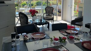 A dining area with a table set for two, a TV on the left, and a view of a garden through large windows.
