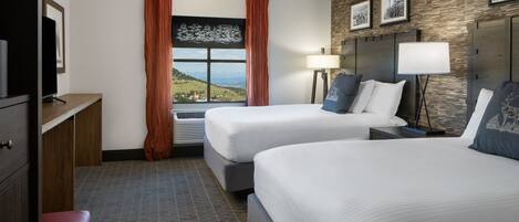 A hotel room with two beds, a desk, a chair, a window with a view, and a lamp.