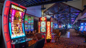 Un casino con máquinas tragamonedas y mesas de juego, alfombra estampada y techo alto con vigas a la vista.