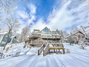 A large, multi-story wooden house with a prominent balcony and a covered deck, surrounded by snow-covered trees and a clear sky.