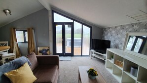 A modern living room with a brown sofa, a coffee table, a TV, and a sliding glass door leading to an outdoor deck. - Log Cabin in the Clwydian Range (Ruthin)