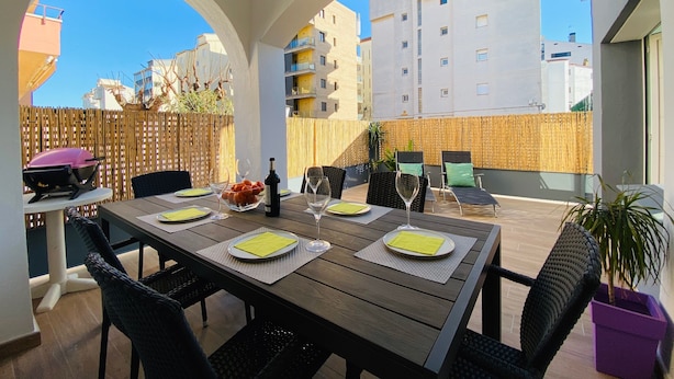Un patio al aire libre con una mesa de madera dispuesta para comer, rodeado de sillas de mimbre y una parrilla.