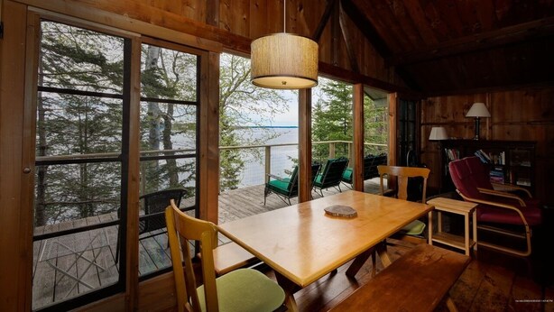 A wooden cabin interior with a large table and chairs, a hanging light fixture, and a view of trees and water through large windows.