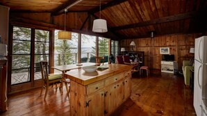 A rustic cabin interior with a wooden dining table, chairs, a kitchen area with a refrigerator, and large windows offering a view of trees.