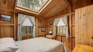 A wooden cabin bedroom with a bed, two windows with curtains, a small table, and a painting on the wall.