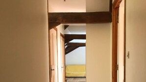 Een smalle gang met houten vloeren en zichtbare balken, die leidt naar een kamer met een geel bed.