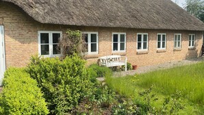 Una casa de ladrillo con techo de paja, ventanas de marco blanco y un pequeño jardín con un banco.