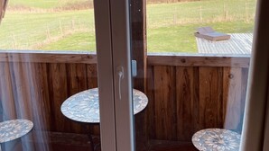 Una habitación con un amplio ventanal que da a una terraza con dos mesas redondas pequeñas y vistas a un campo cubierto de césped.