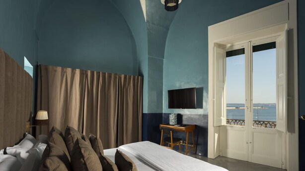 A bedroom with a bed, curtains, a desk with a chair, a TV, and a view of the sea through a door. - Palazzo Colombo (Gallipoli)