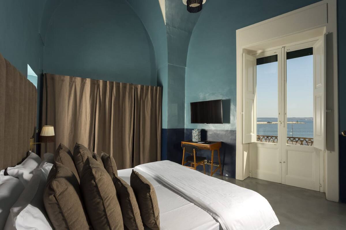 A bedroom with a bed, curtains, a desk with a chair, a TV, and a view of the sea through a door.