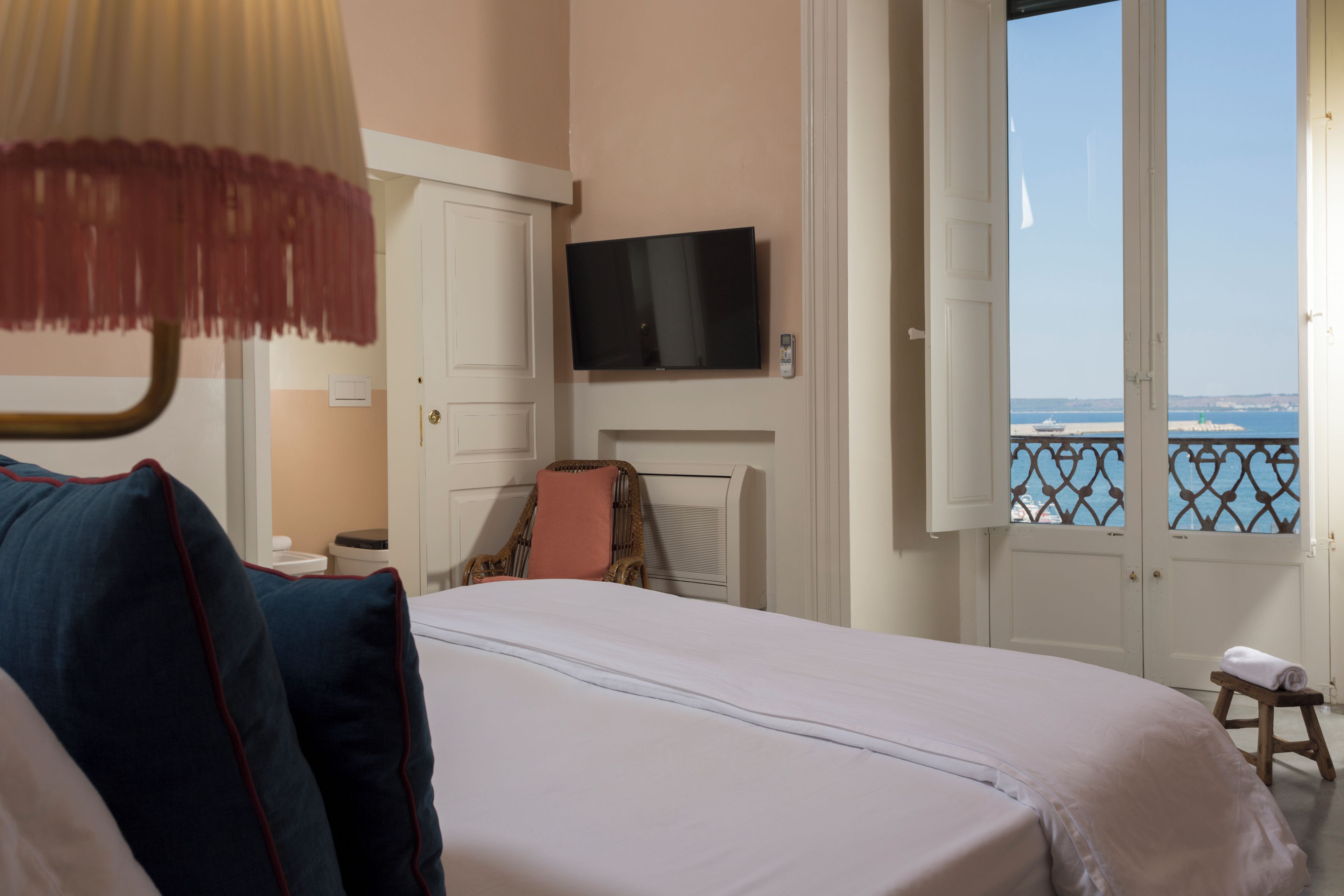 Une chambre d’hôtel avec un lit, un téléviseur fixé au mur, une fenêtre donnant sur la mer et un petit tabouret près de la fenêtre.
