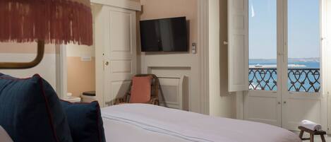 Une chambre d’hôtel avec un lit, un téléviseur fixé au mur, une fenêtre donnant sur la mer et un petit tabouret près de la fenêtre.