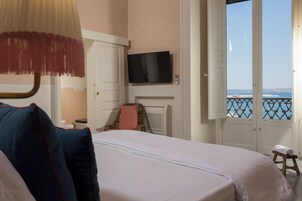 Una habitación de hotel con cama, televisor montado en la pared, ventana con vista al mar y un taburete cerca de la ventana.