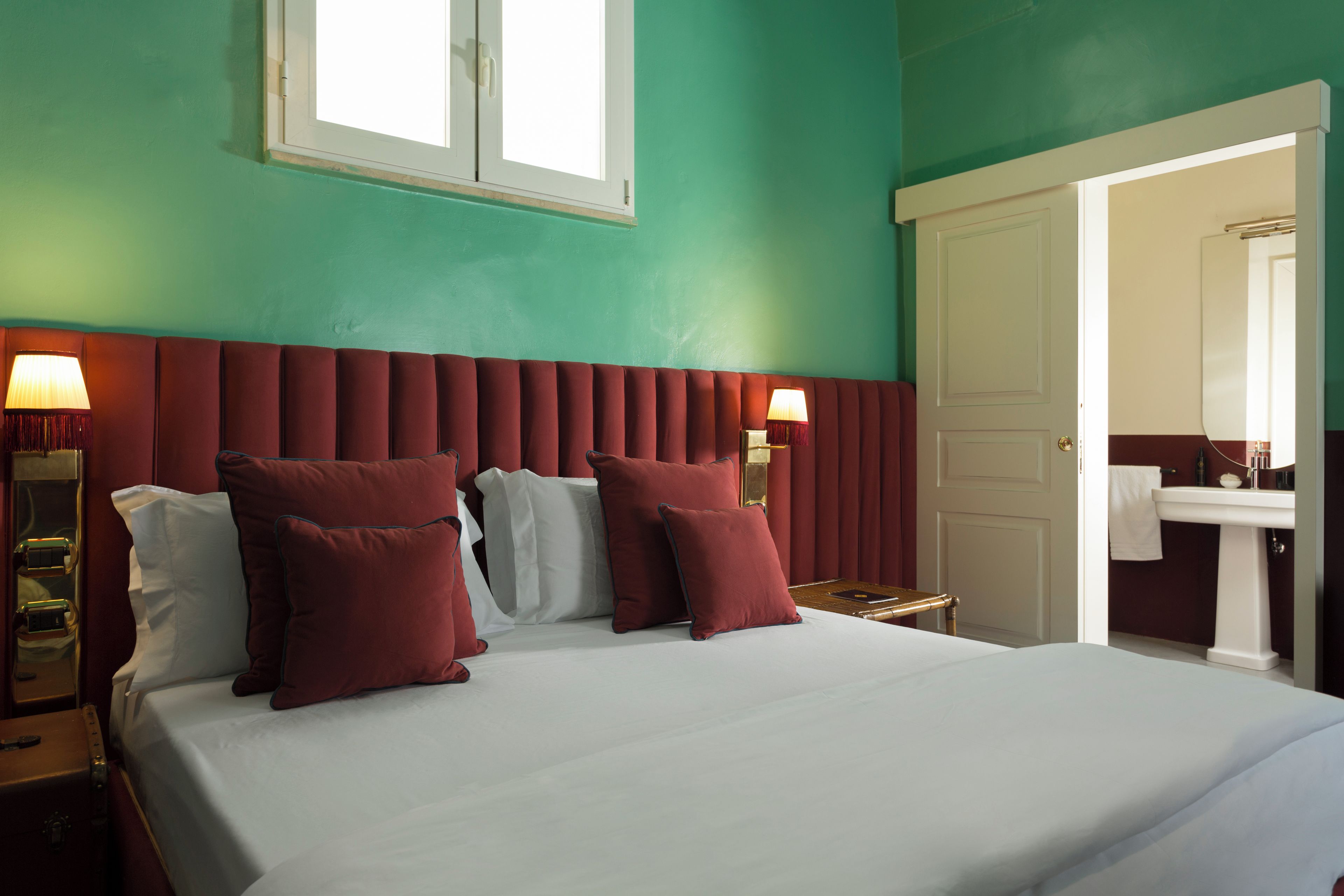 A hotel room with a large bed, two bedside lamps, a window, and a bathroom visible through an open door.