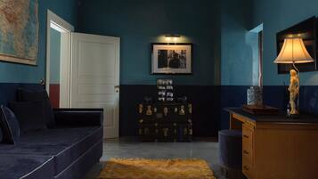 A room with a dark blue wall, a white door, a blue sofa, a wooden dresser, a lamp, and a framed picture.