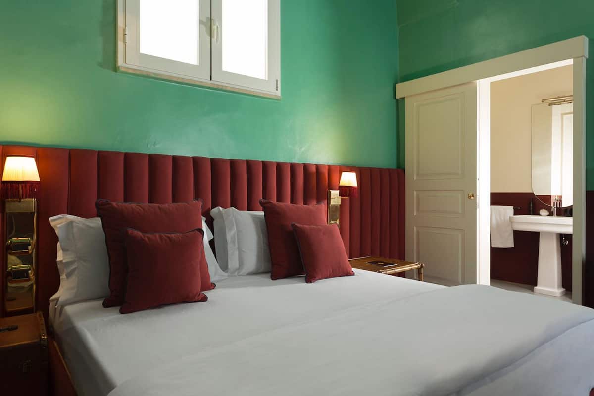 A hotel room with a bed, two bedside lamps, a nightstand, a window, and a bathroom with a sink and mirror.